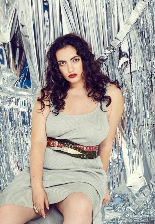 Portrait of an adult woman with dark curly hair and red lipstick, wearing a dress with a belt, seated against a shiny silver background.