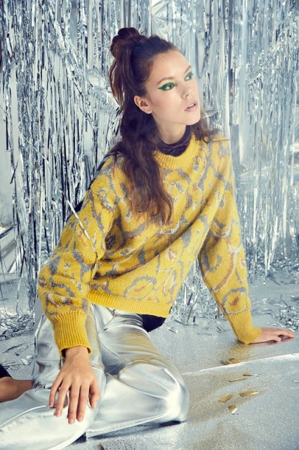 A woman with an updo and green eyeliner poses while sitting on a silver reflective surface, wearing a yellow patterned sweater and silver pants.