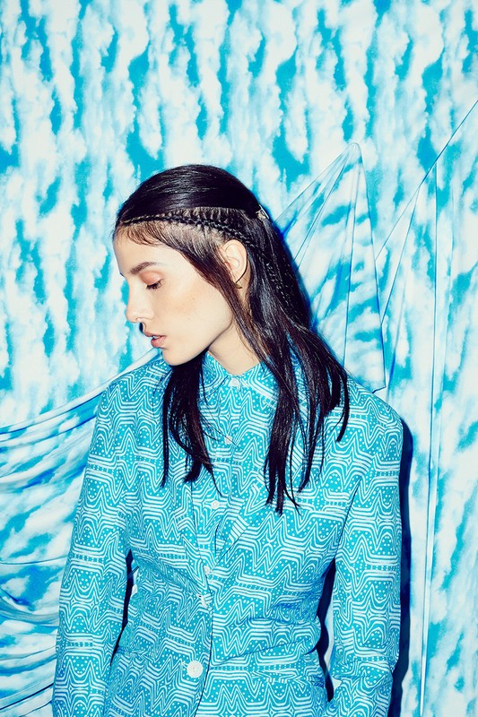 Brunette woman in a patterned teal suit poses in front of a matching patterned teal and white background, with her head angled down.