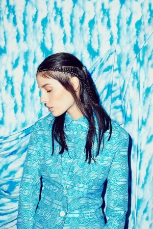 Brunette woman in a patterned teal suit poses in front of a matching patterned teal and white background, with her head angled down.