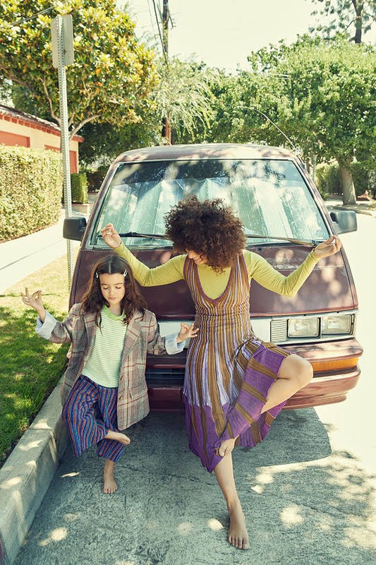 Mother and daughter doing yoga poses in front of a vintage minivan on a sunny street, wearing casual, colorful clothing.