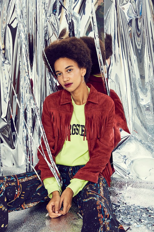 A young woman with an afro sits in front of a reflective silver curtain wearing a red jacket, yellow shirt, and patterned pants.