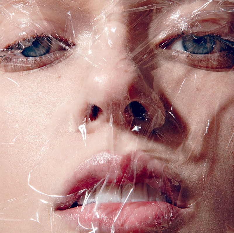 Close-up portrait of a person's face covered in clear plastic wrap, highlighting their eyes, nose, and mouth with soft, diffused lighting.