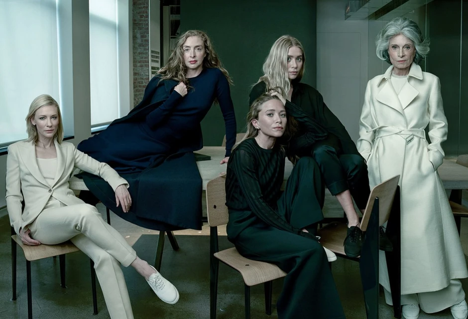 Group portrait of five women in a modern interior setting, wearing tailored clothing in black, white, and blue, seated and standing.