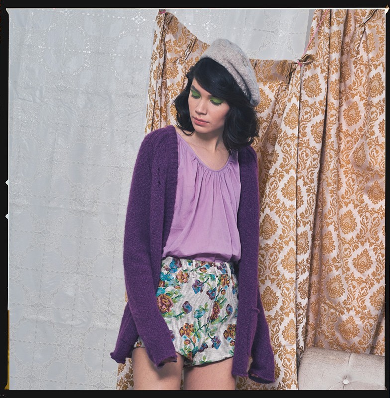 Woman in floral shorts, purple cardigan, and beret poses against a patterned backdrop, showcasing a blend of casual and vintage fashion.