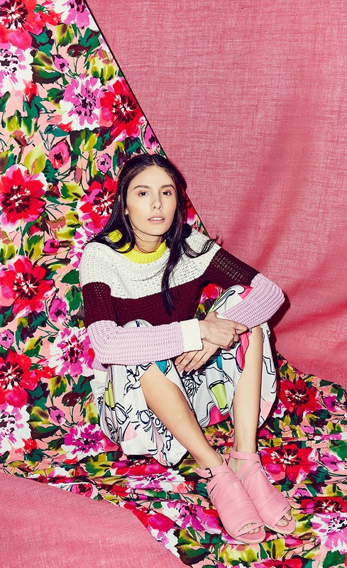 Young woman in colourful sweater and skirt posing on a floral backdrop with pink cloth, looking at the camera indoors in studio lighting.