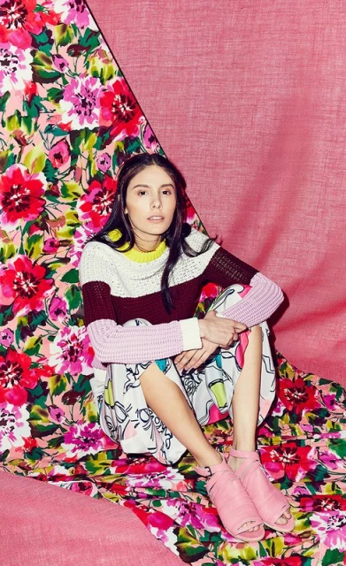 Young woman in colourful sweater and skirt posing on a floral backdrop with pink cloth, looking at the camera indoors in studio lighting.