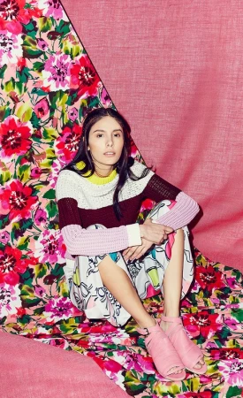 Young woman in colourful sweater and skirt posing on a floral backdrop with pink cloth, looking at the camera indoors in studio lighting.