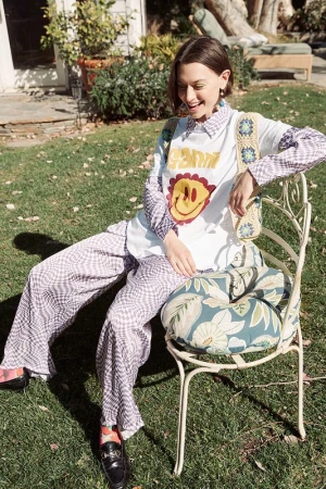 A young woman sits in a garden chair wearing loose patterned pants and a t-shirt with a smiley face graphic, bathed in sunlight.