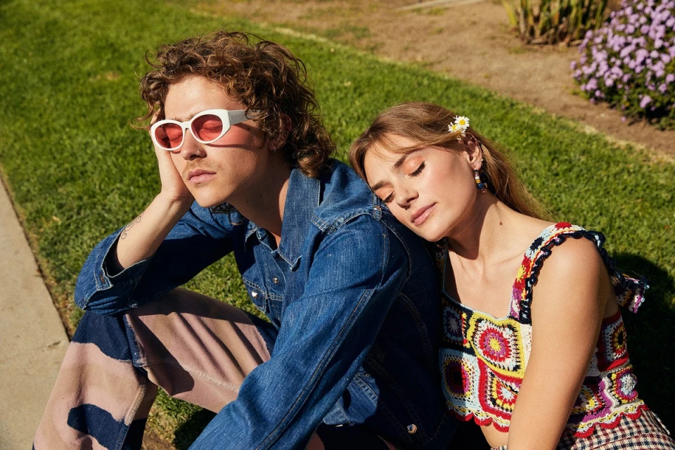 Young couple sitting outside on the grass in the sunlight; the woman is leaning her head on the man's shoulder.