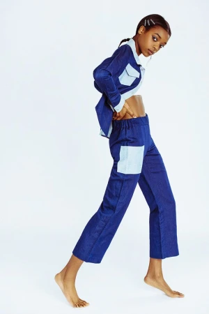 A young Black woman posing in a matching blue denim jacket and trousers against a clean white seamless studio background.