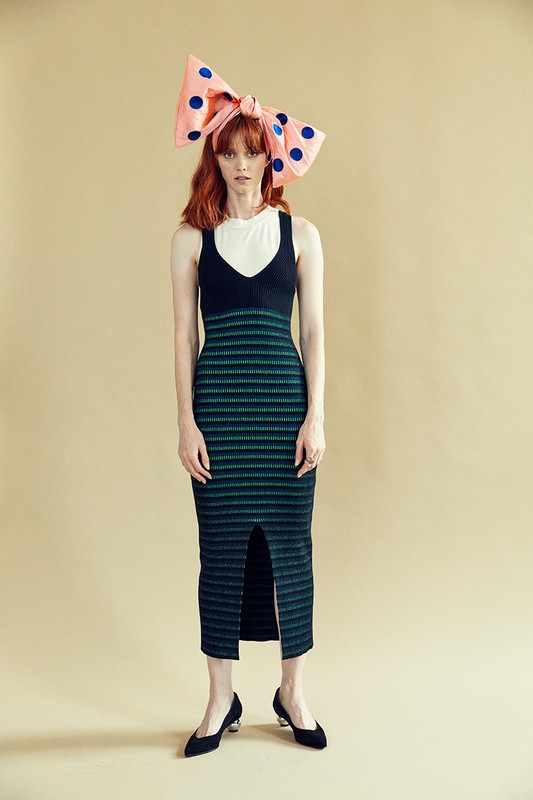Model with red hair wearing a large pink bow with blue polka dots, a black and green striped dress, and black shoes.