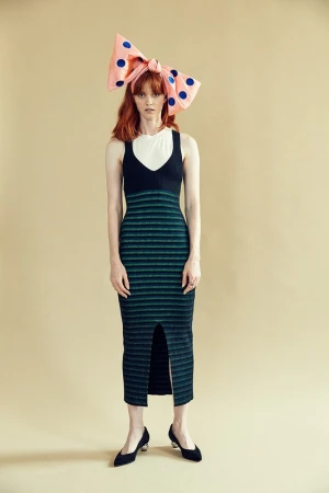 Model with red hair wearing a large pink bow with blue polka dots, a black and green striped dress, and black shoes.