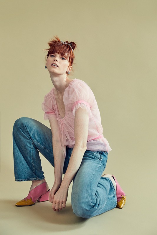 Redhead woman in pink top and jeans, squatting pose against pale yellow backdrop in a fashion-style photograph.  Stylish and vibrant.