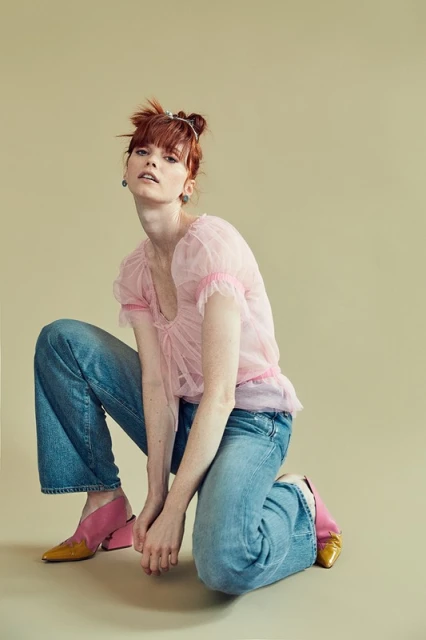 Redhead woman in pink top and jeans, squatting pose against pale yellow backdrop in a fashion-style photograph.  Stylish and vibrant.