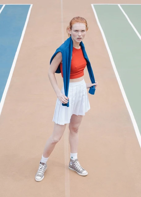 A woman with red hair in athletic wear, including a white skirt and red shirt, stands on a tennis court with blue and green accents.