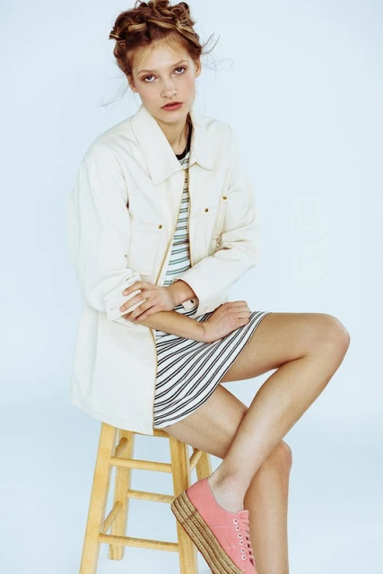 Young woman with braided hair sits on a wooden stool in a photo studio wearing a striped dress, white jacket, and pink platform shoes.