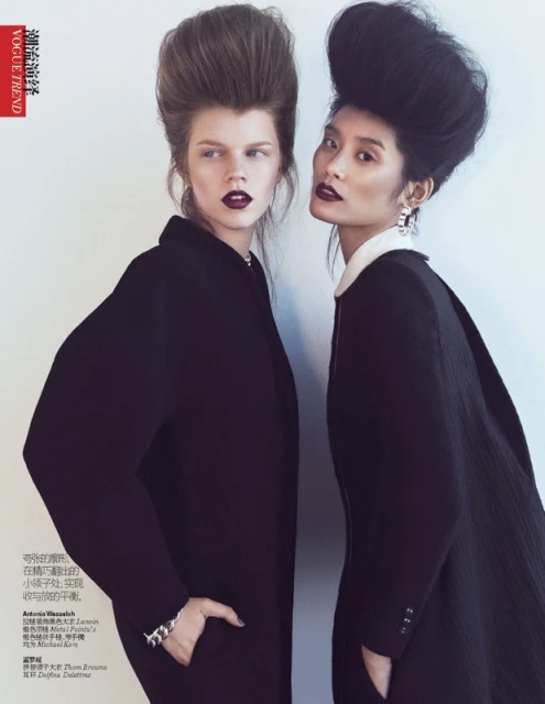 Two fashion models pose in black coats with high-volume hairstyles and dark lipstick against a neutral background in a studio setting.