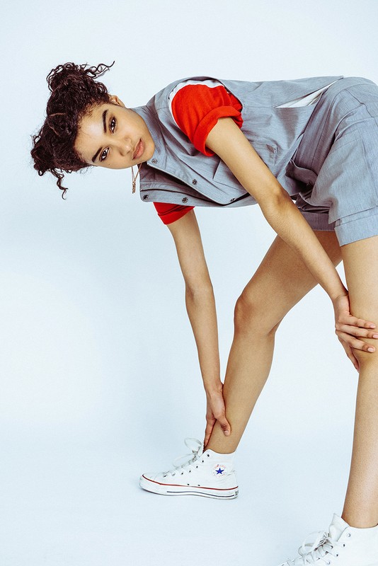 Young adult woman in streetwear fashion poses with a bend wearing a red t-shirt, grey vest, and shorts in a white studio setting.