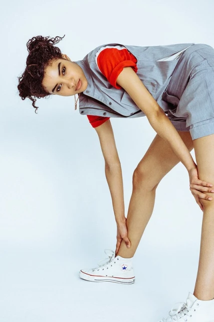 Young adult woman in streetwear fashion poses with a bend wearing a red t-shirt, grey vest, and shorts in a white studio setting.