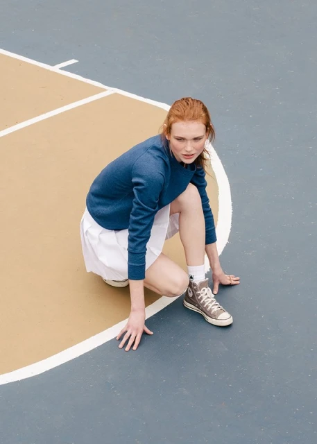Young adult woman with red hair in a navy sweater and white tennis skirt poses on a blue and beige outdoor court with sneakers.