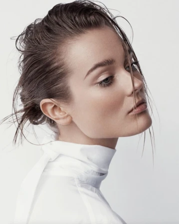 Close-up profile portrait of a woman with wet hair styled in a bun, wearing a white high-neck top against a seamless white background.