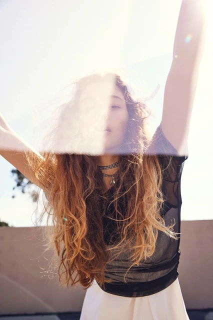Woman in black mesh top with long wavy hair, white skirt, and necklace stands with arms raised against a bright sunny sky.