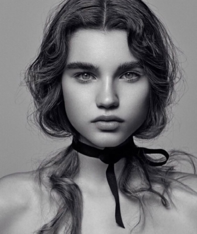 Close-up monochrome portrait of a young woman with wavy hair wearing a black ribbon choker necklace against a grey backdrop.