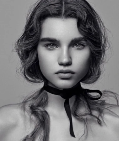 Close-up monochrome portrait of a young woman with wavy hair wearing a black ribbon choker necklace against a grey backdrop.