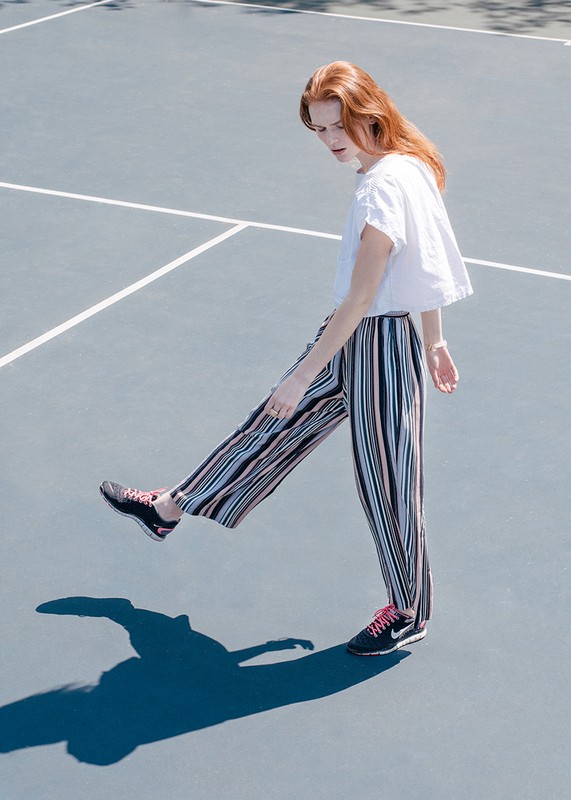A stylish red-haired woman poses in vertically striped pants and sneakers on a blue outdoor surface with hard shadows.
