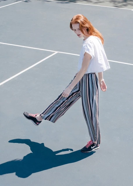 A stylish red-haired woman poses in vertically striped pants and sneakers on a blue outdoor surface with hard shadows.