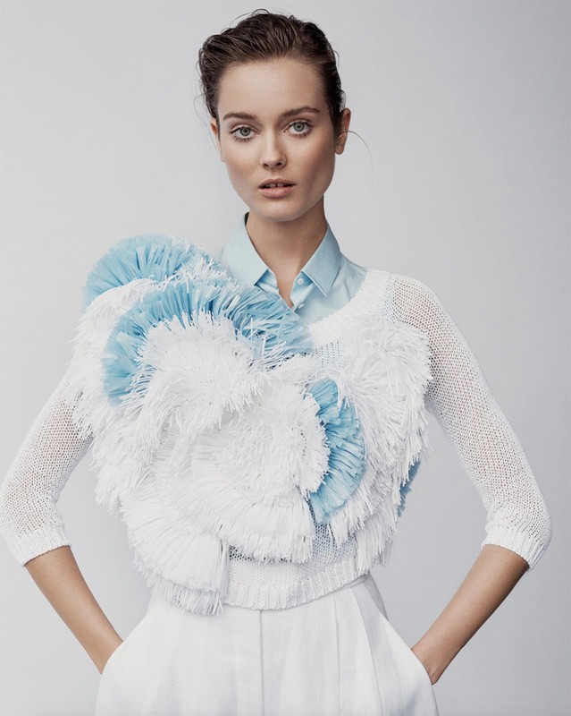 Fashion portrait of a young adult female model wearing a white and light-blue textured top and white pants against a light-grey backdrop.