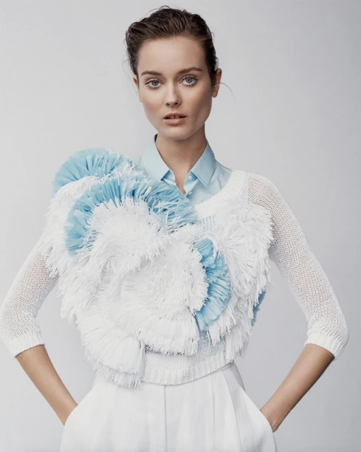 Fashion portrait of a young adult female model wearing a white and light-blue textured top and white pants against a light-grey backdrop.