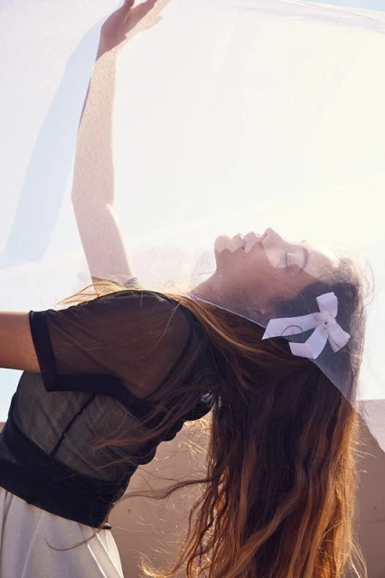 Woman in black top and white skirt with hair bow, arm raised under white translucent fabric in outdoor setting with sunlight. 