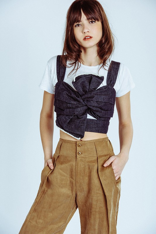 Fashion portrait of a young woman in a white t-shirt, tied dark blue top, and tan pants, striking a confident pose in a studio setting.