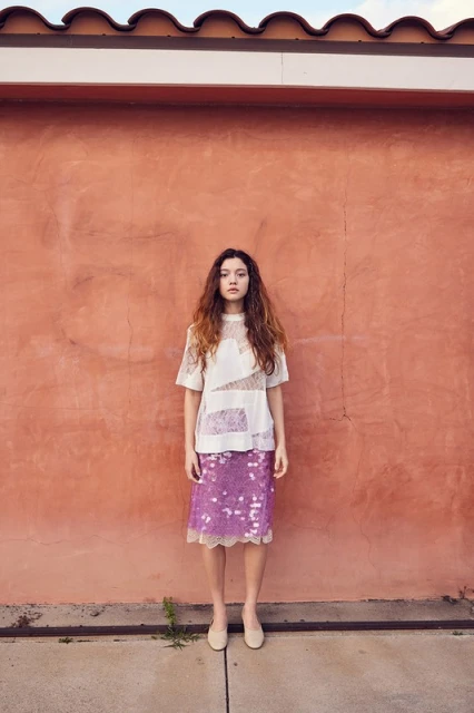 Young woman in a white shirt and purple sequined skirt stands against an orange textured wall outdoors on a sunny day.