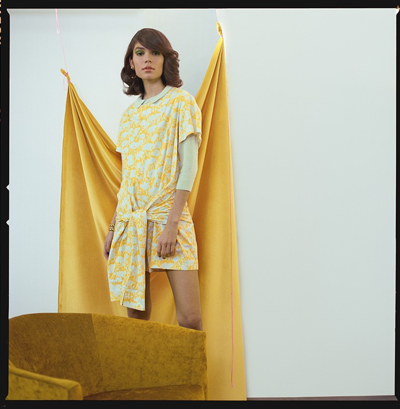 Adult woman poses in a yellow and white patterned dress indoors in front of a yellow drape and beside a vintage yellow couch.