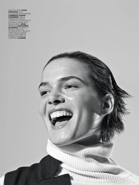 Black and white portrait of a smiling woman with wet-look hair wearing a turtleneck and vest in a studio with soft, diffused lighting.