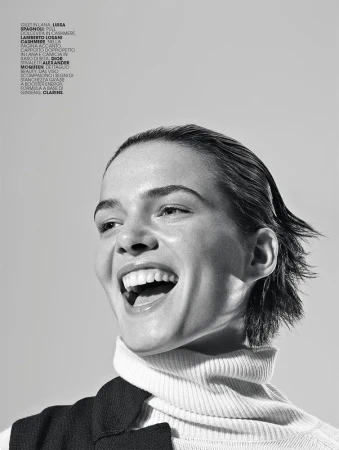 Black and white portrait of a smiling woman with wet-look hair wearing a turtleneck and vest in a studio with soft, diffused lighting.