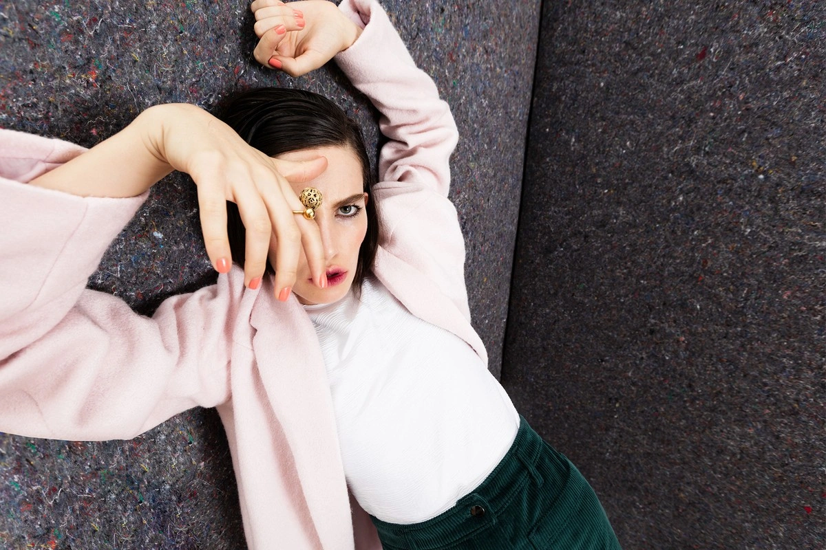 Woman wearing a pale pink coat and green corduroy trousers reclining against a textured grey wall