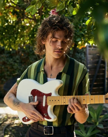 Young man with curly brown hair playing a white electric guitar in an outdoor garden setting in Los Angeles.