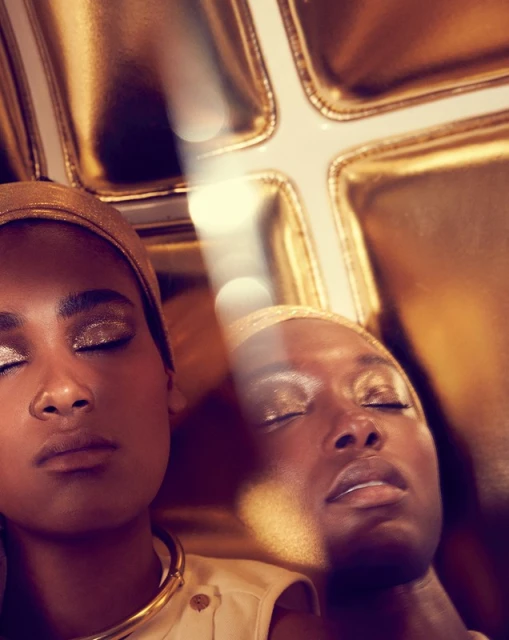 Golden hour portrait of two Black women with glitter eyeshadow wearing headscarves and gold jewelry, with a bright light.