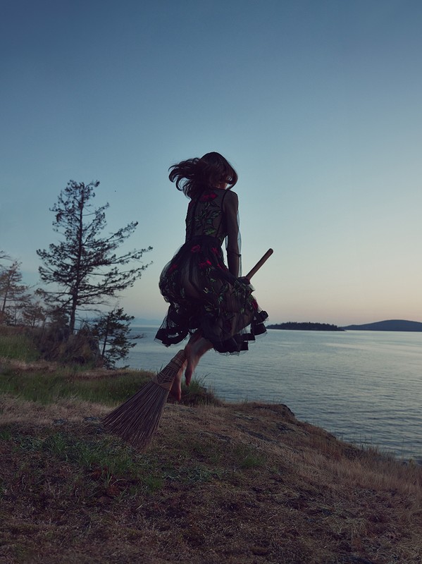 Woman in dark dress with floral accents jumps off a grassy cliff on a broom at dusk, water and trees in the background.
