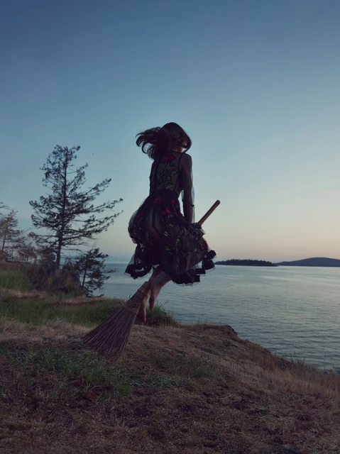 Woman in dark dress with floral accents jumps off a grassy cliff on a broom at dusk, water and trees in the background.