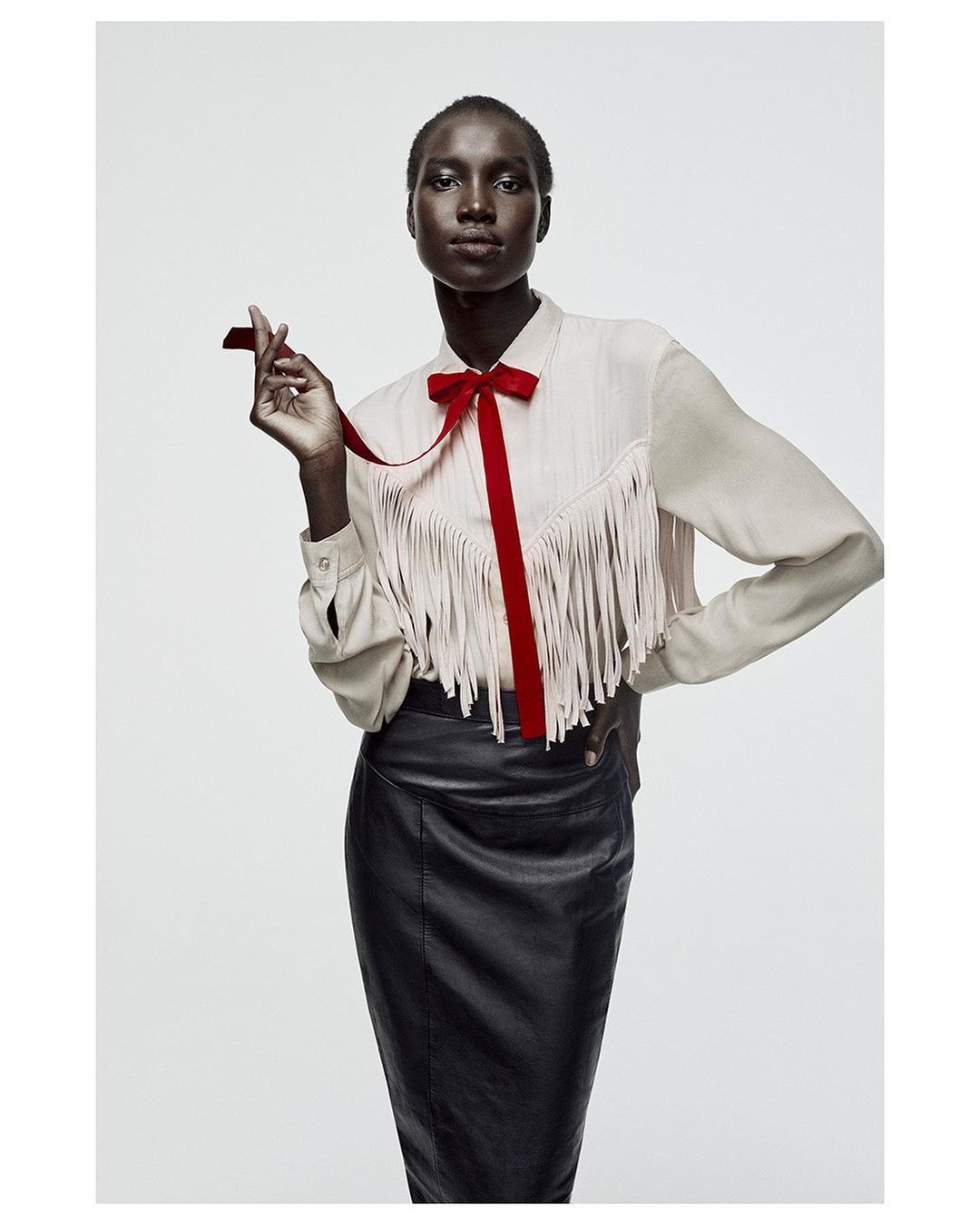 Studio shot of a Black woman in a cream blouse with fringe and a red bow, paired with a black leather skirt against a white backdrop.
