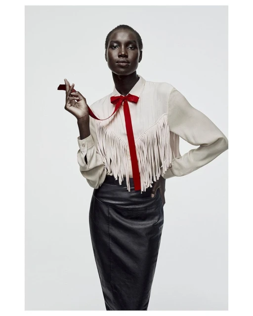 Studio shot of a Black woman in a cream blouse with fringe and a red bow, paired with a black leather skirt against a white backdrop.
