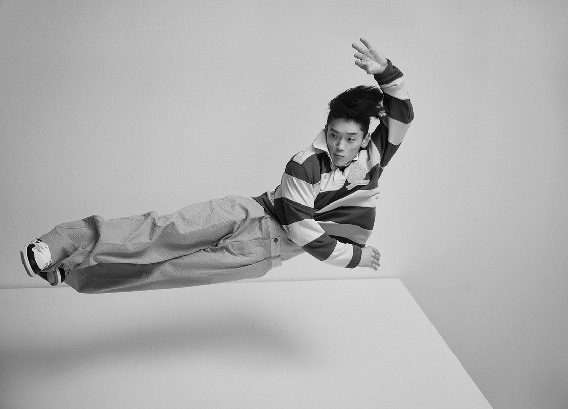 Asian man in mid-air with striped shirt and textured pants, jumping or leaping over a white block in a monochrome indoor setting.