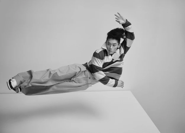 Asian man in mid-air with striped shirt and textured pants, jumping or leaping over a white block in a monochrome indoor setting.