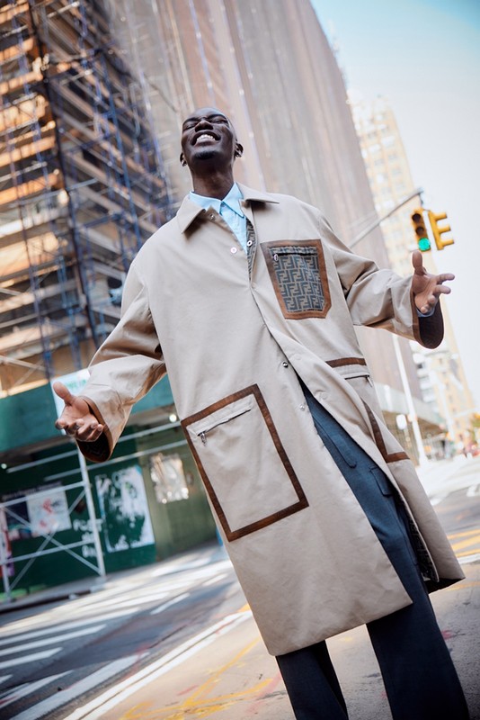 An African American man in an open tan trench coat stands on a city street, smiling against a backdrop of buildings and scaffolding.