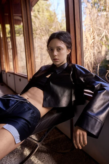 A young woman in a black leather jacket and shorts sits in a chair near a window with an outdoor view, posing for a fashion shot.
